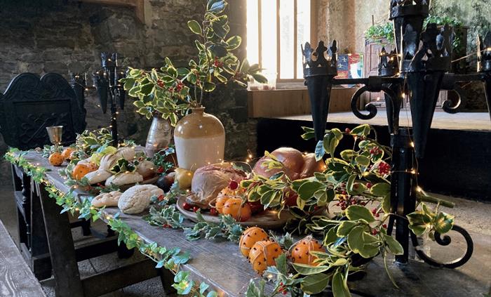 A long wooden table laden with a festive spread of bread, oranges and a roasted fowl, decorated with holly branches and fairy lights.